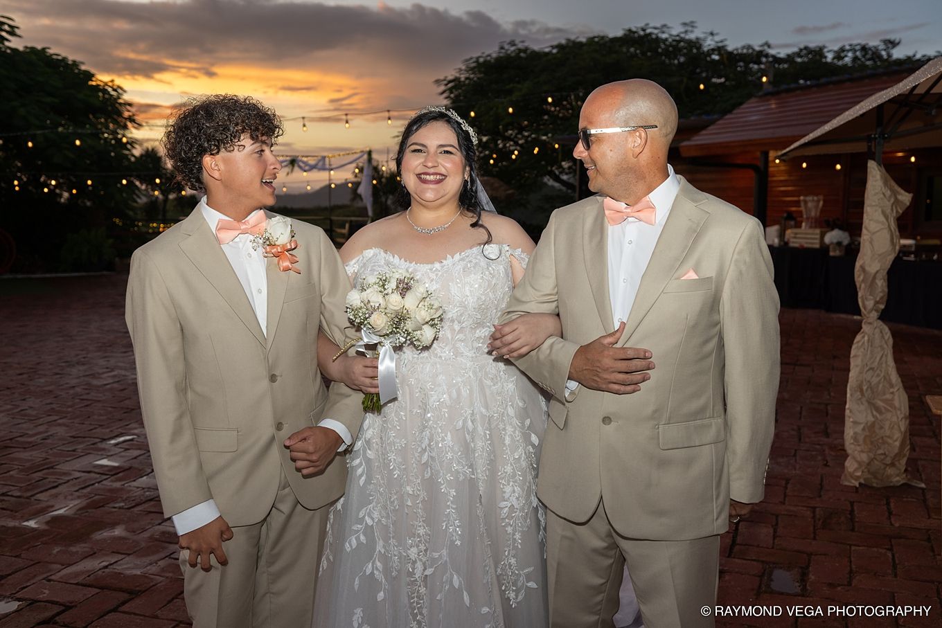 Bride in white gown holds bouquet flanked by two men in tan suits at outdoor wedding reception at sunset.