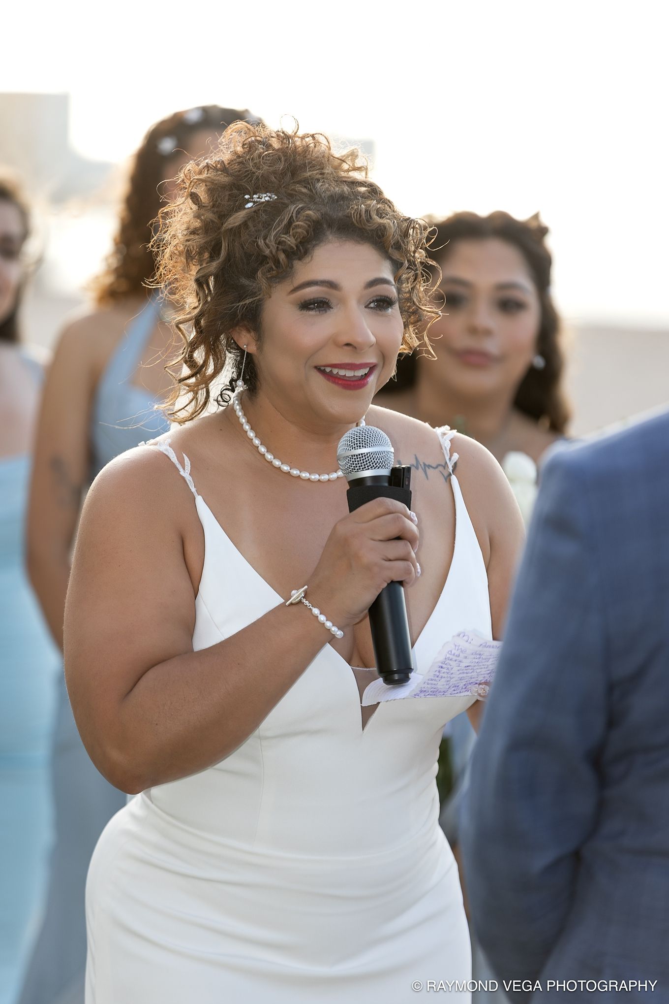 Bride in white dress holding microphone speaks at outdoor wedding ceremony with bridesmaids in blue dresses behind her.