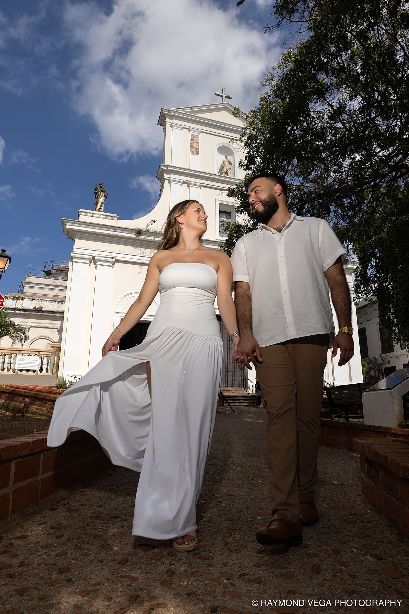 Couple in white attire holding hands near a white church under blue skies, captured by Raymond Vega Photography. Old San Juan  Puerto Rico