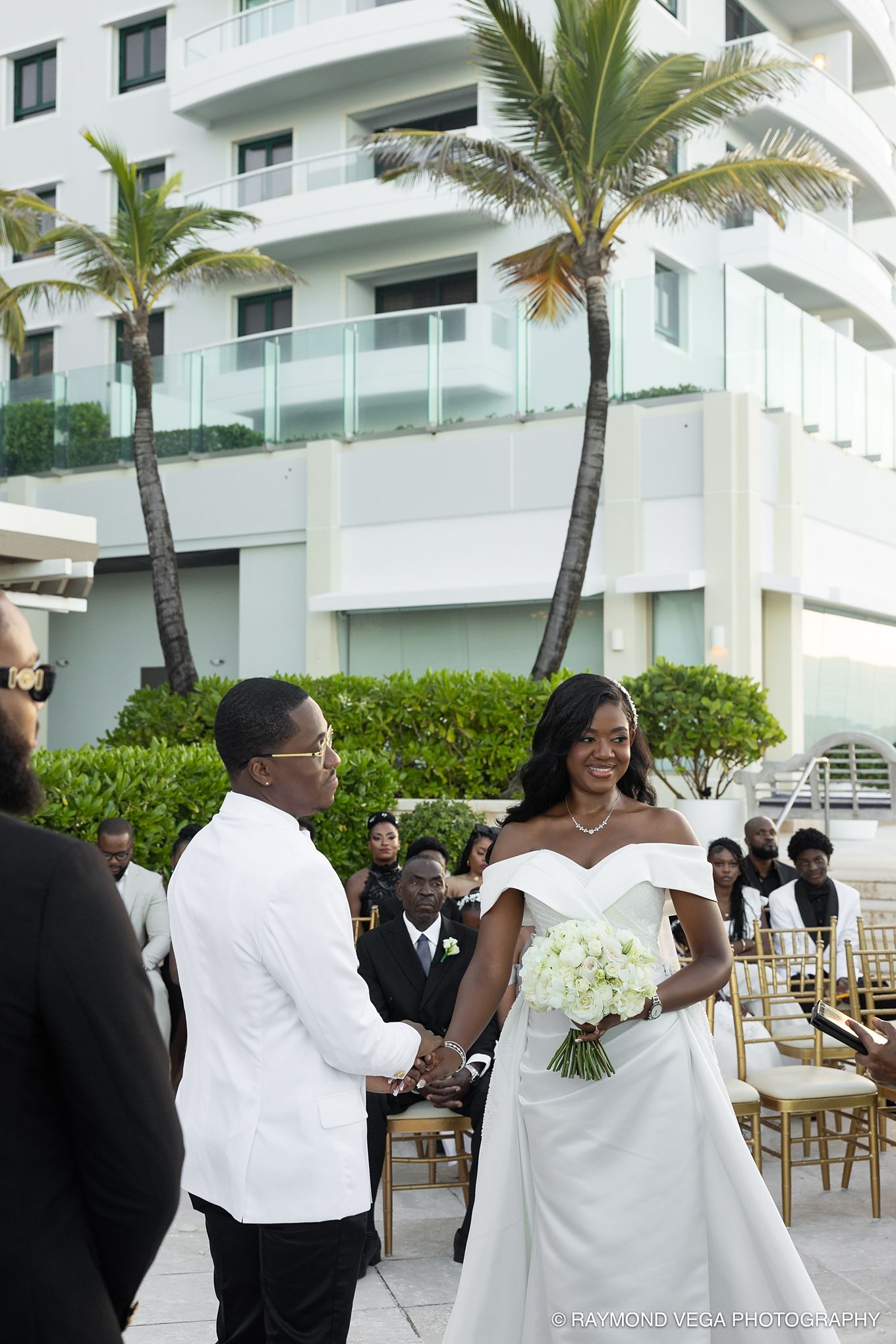 Bride and groom holding hands during outdoor wedding ceremony at beachside resort with palm trees.