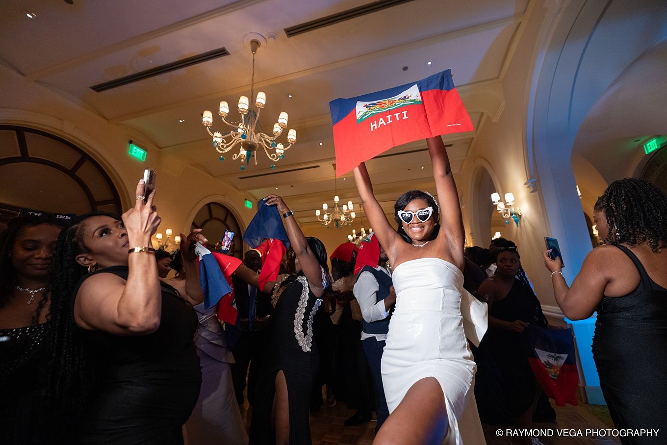 Woman in white dress holding Haitian flag celebrates at elegant wedding reception with guests dancing in ballroom.