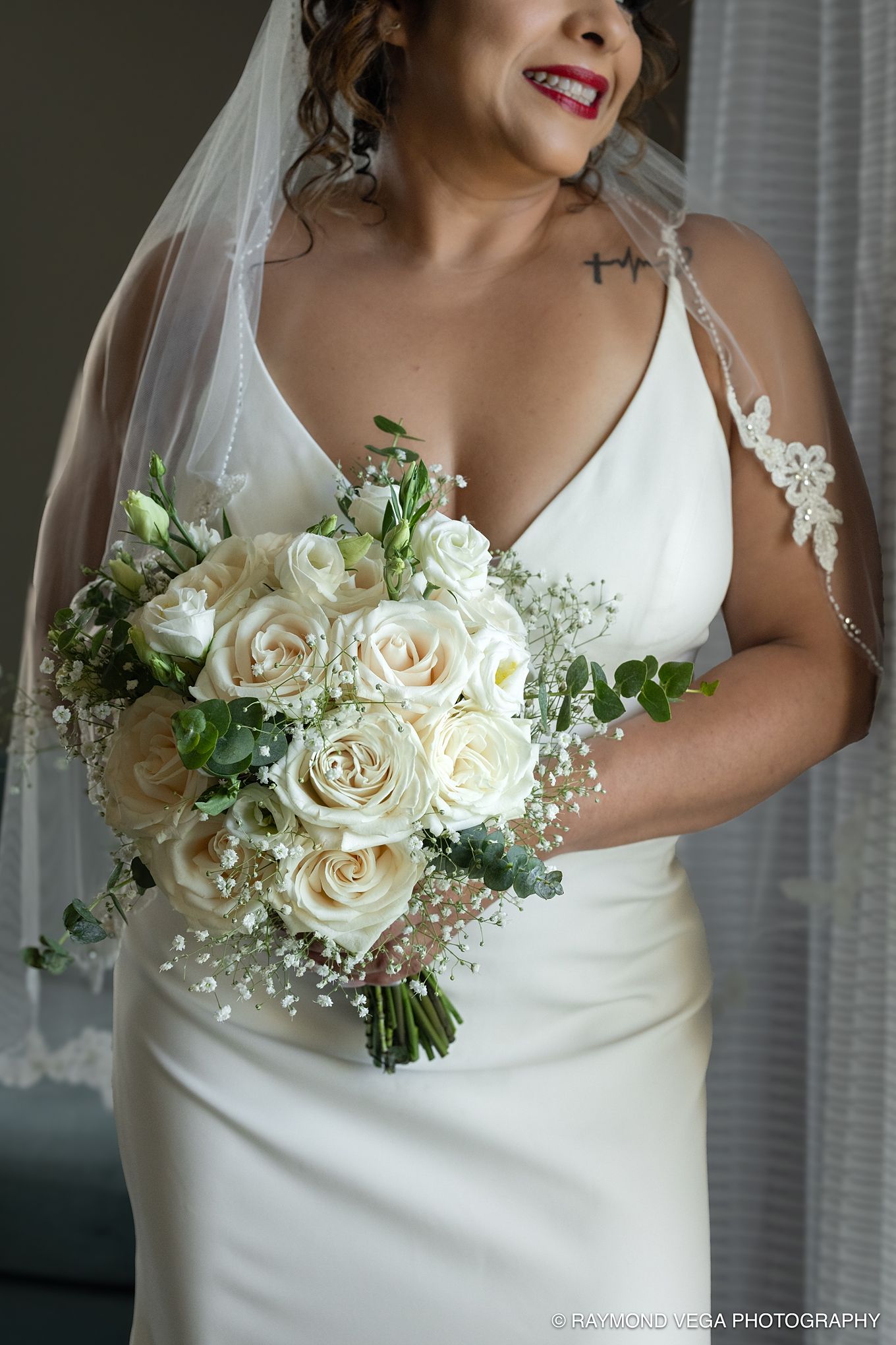 Bride in white gown holding elegant bouquet of ivory roses and baby's breath, wearing lace veil on wedding day.