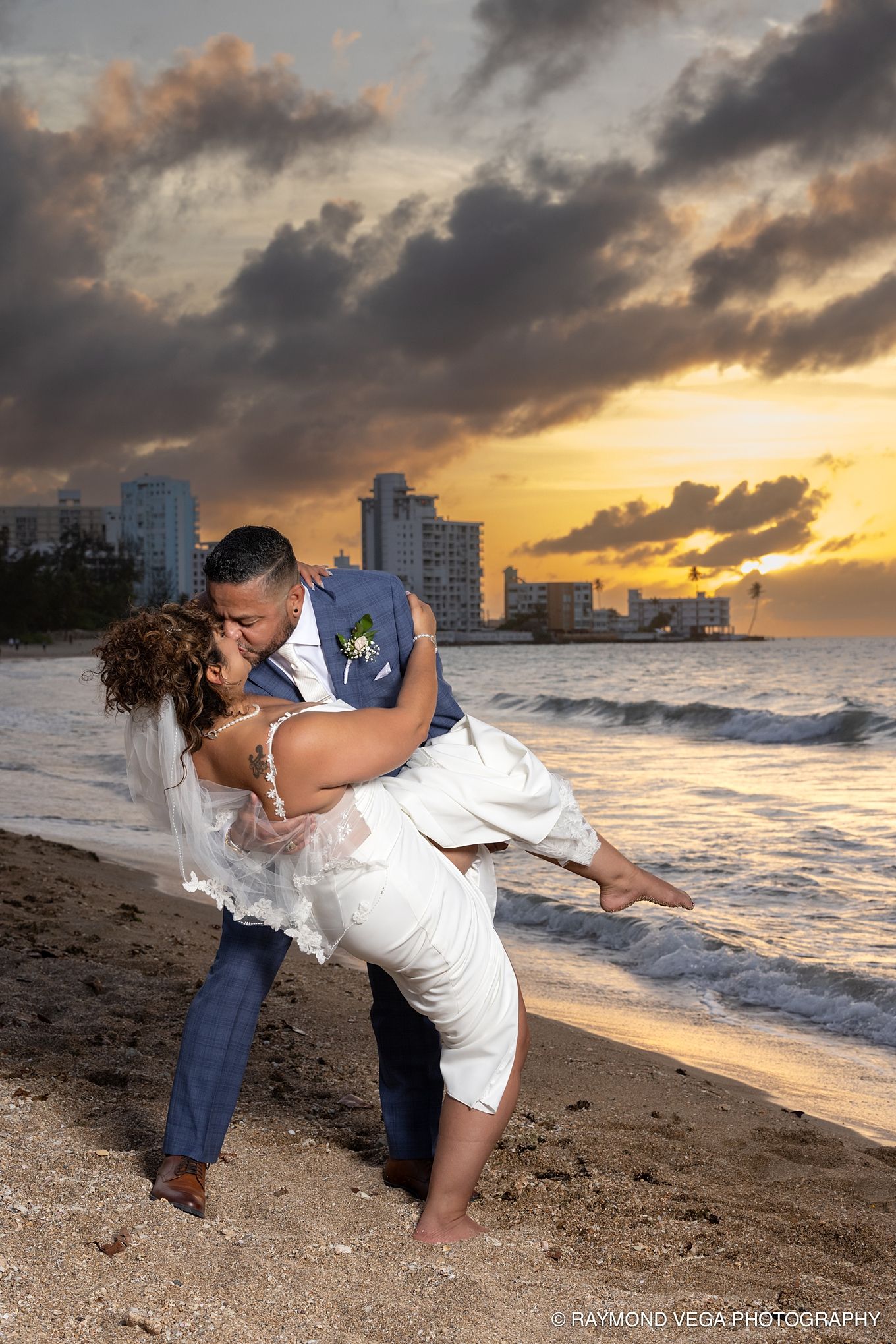 Couple shares romantic dip kiss on beach at sunset with city skyline backdrop during wedding celebration.