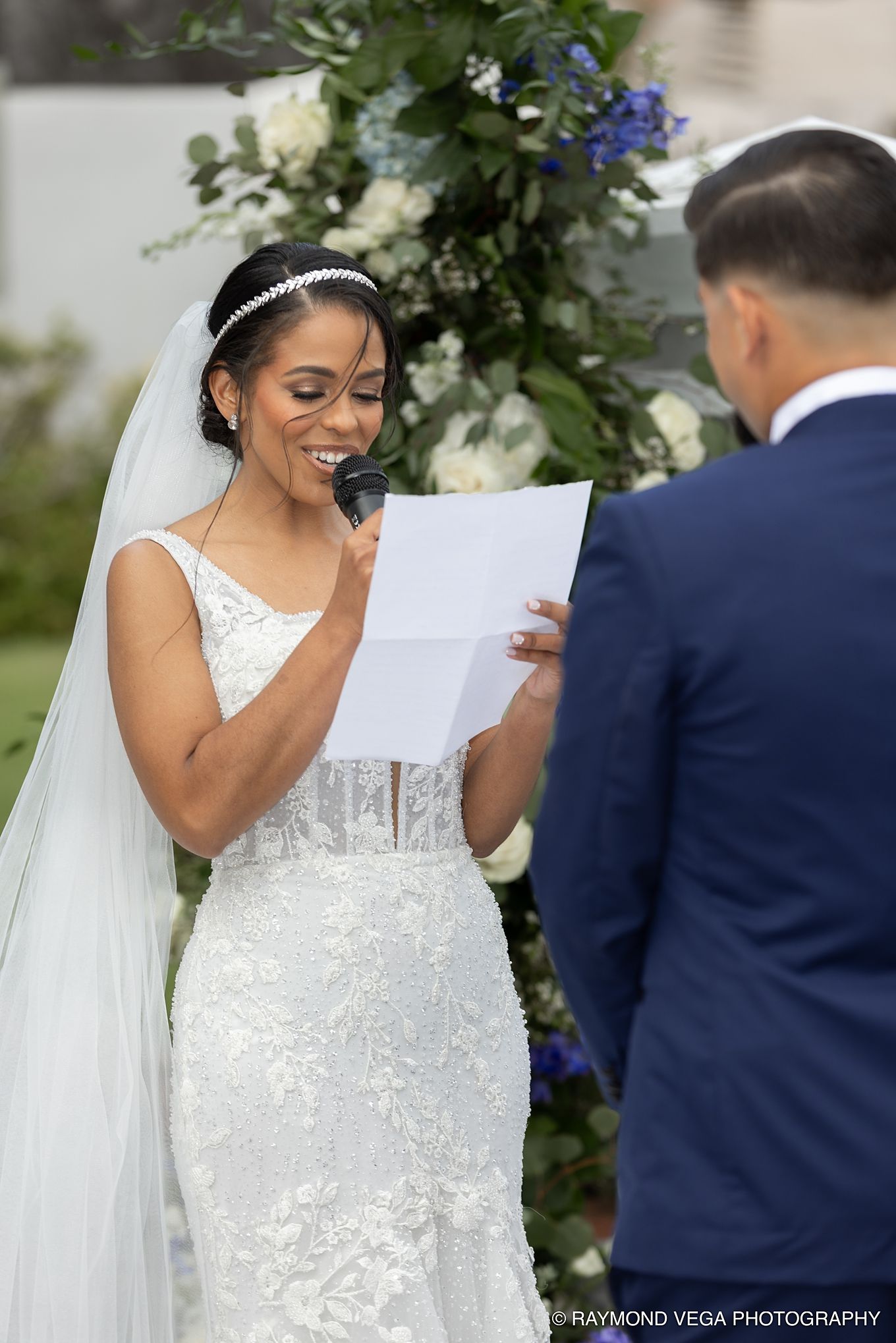 Smiling bride reads wedding vows from paper at outdoor ceremony, wearing lace gown and veil with floral arch backdrop. Antiguo Casino Puerto