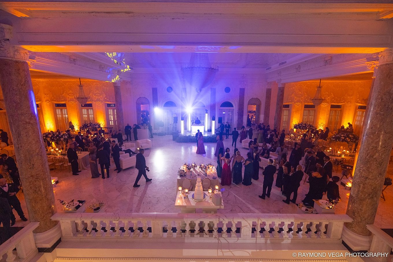 Elegant wedding reception in a grand ballroom with warm amber uplighting, marble columns, and guests dancing on the floor.Antiguo Casino Pue