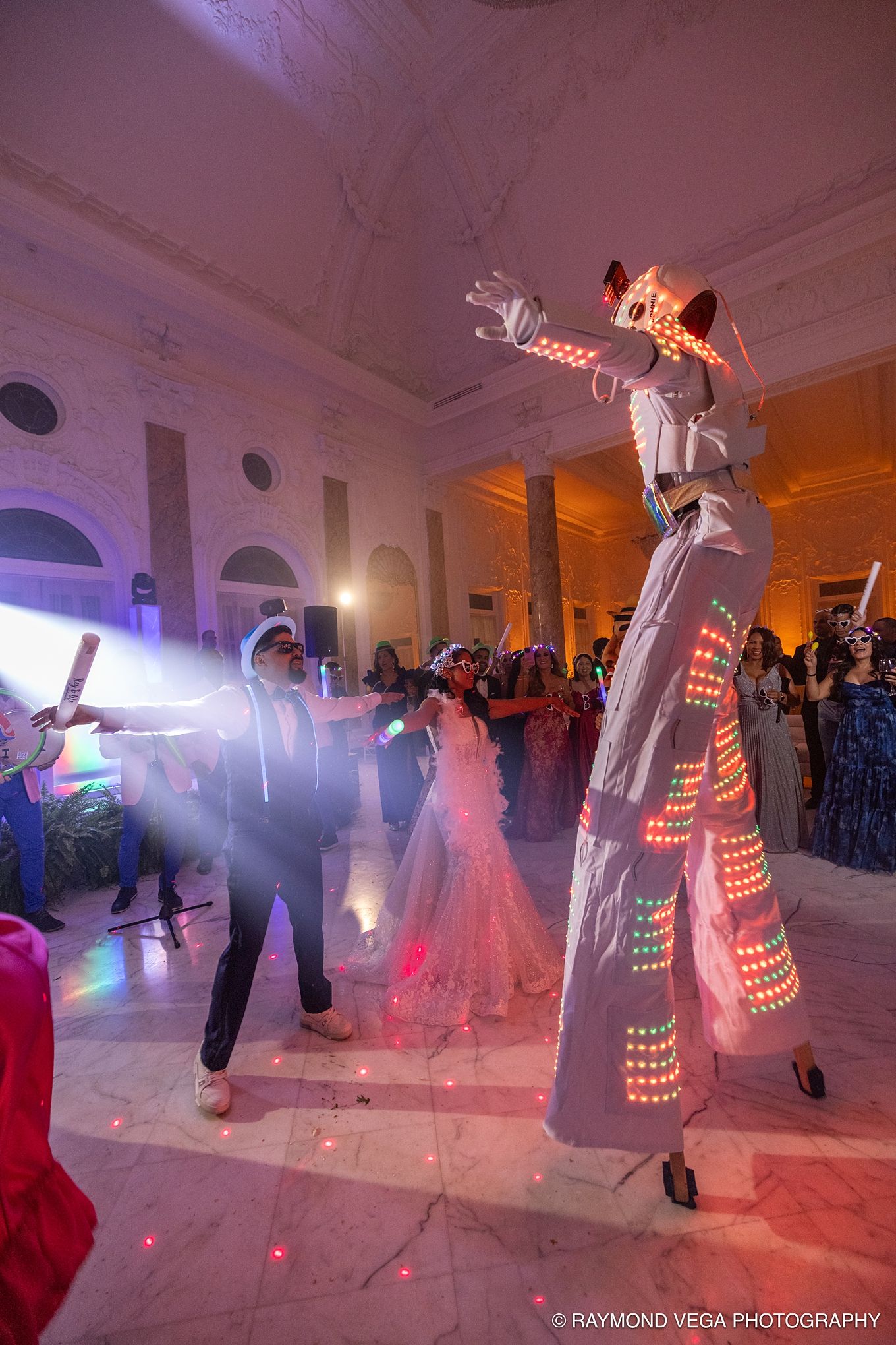 A stilt walker in LED costume performs at an elegant wedding reception with glowing lights and festive crowd. Sak Entretaiment