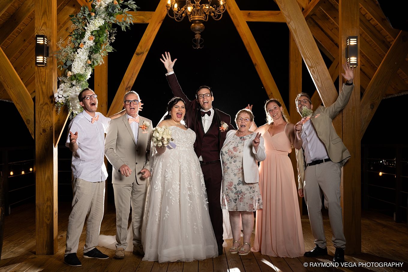 Wedding party celebrating with bride and groom under floral arch in rustic barn venue at night.