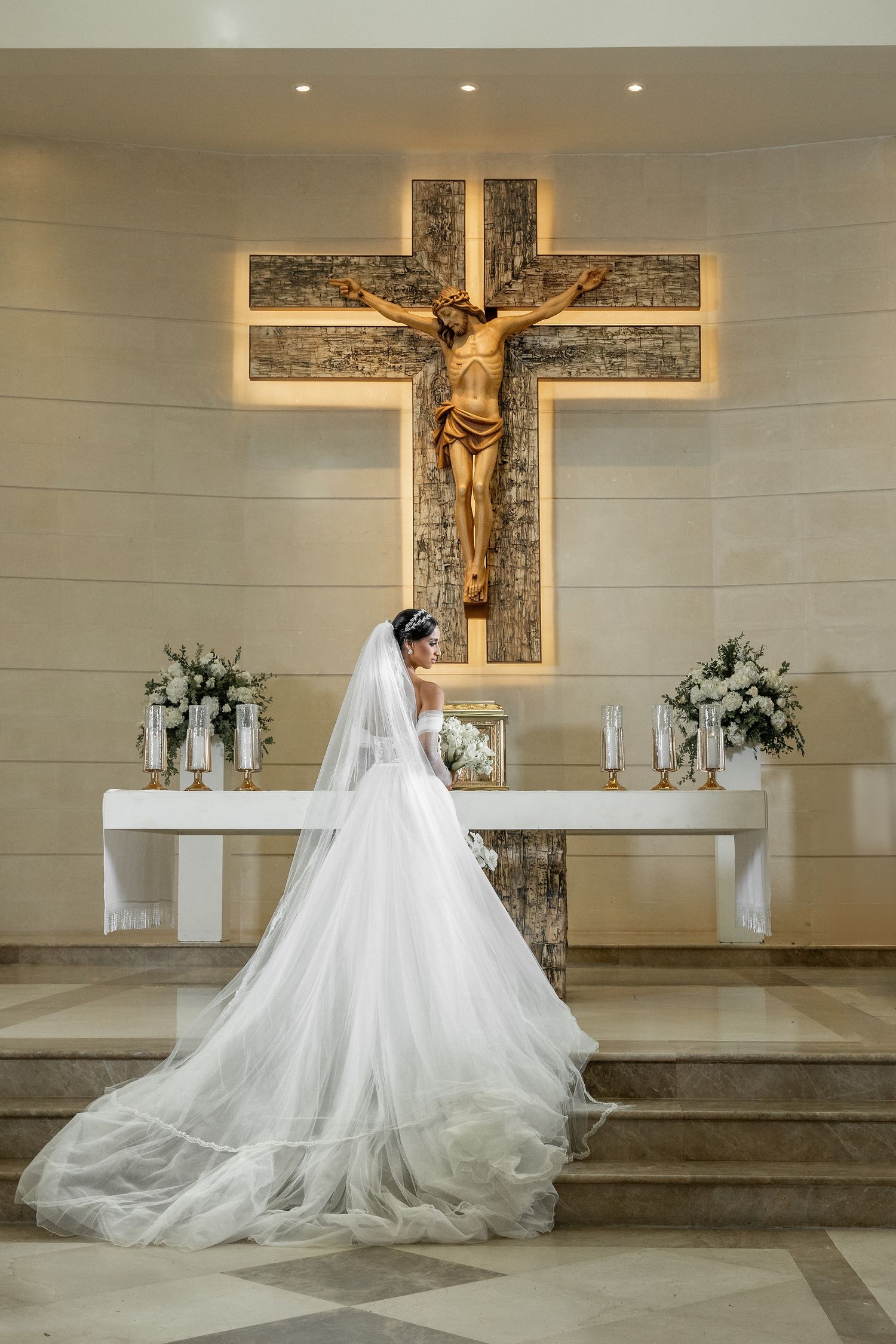 Bride in white gown and veil stands before lit crucifix altar in elegant church wedding ceremony.