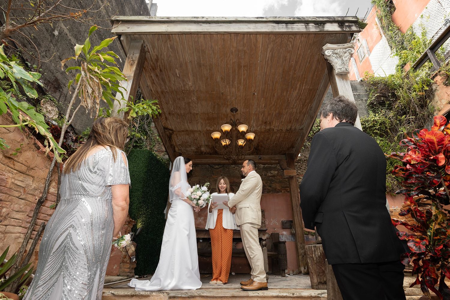 Bride and groom exchange vows with officiant under rustic outdoor pergola surrounded by lush greenery at intimate wedding ceremony.
