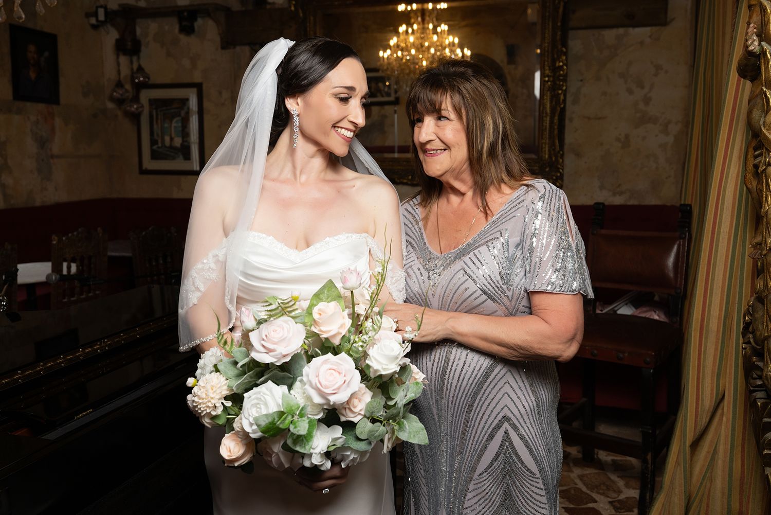 Bride in white gown and veil holding pink rose bouquet smiling with older woman in silver dress at wedding venue.
