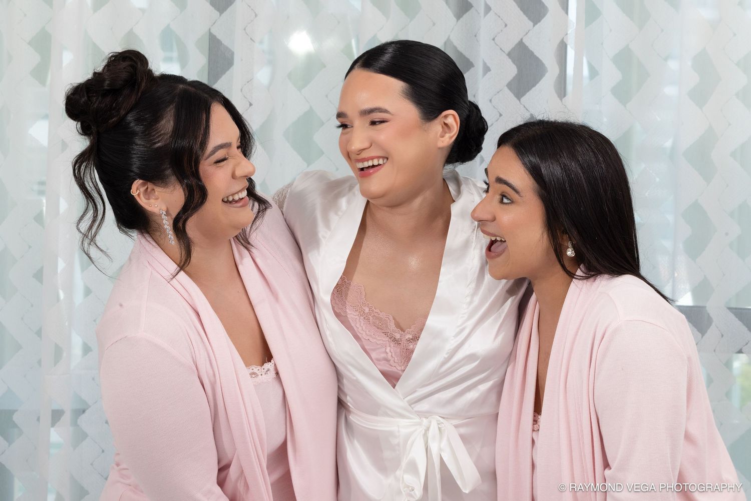 Three women in pink robes laughing and embracing together in a bright, elegant bathroom setting.