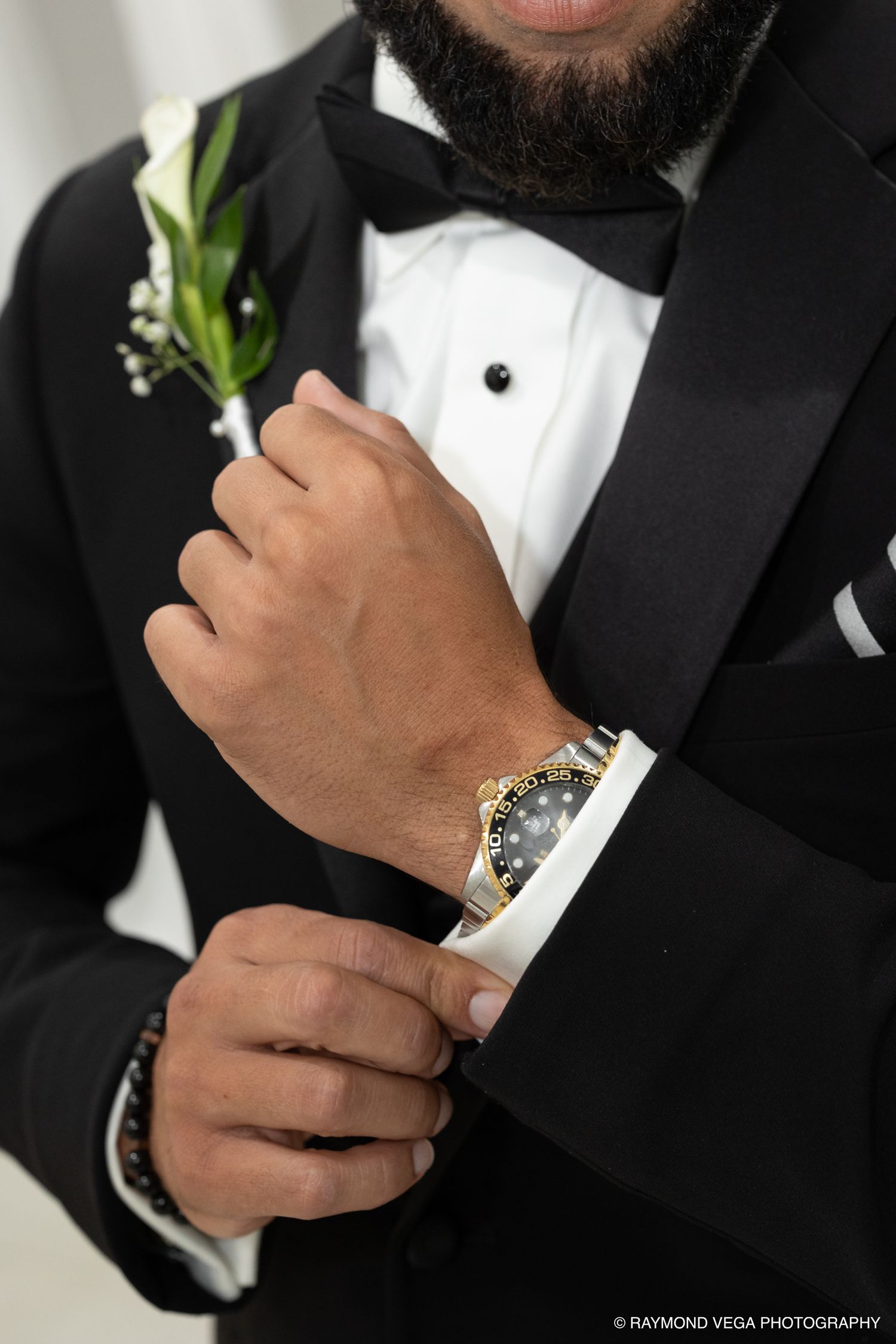 Groom in black tuxedo adjusting cufflinks wearing gold Rolex watch with white calla lily boutonniere.