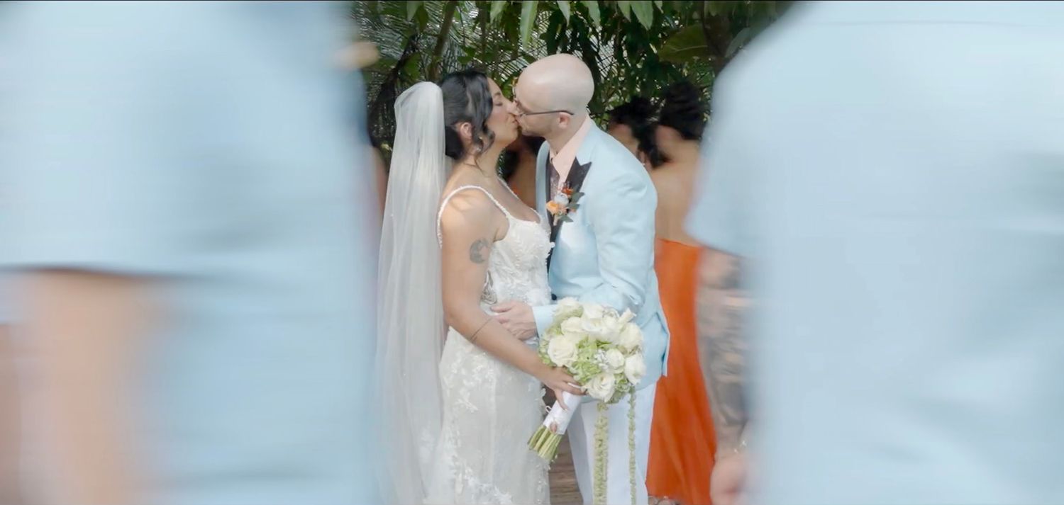 Bride and groom share a kiss at the altar surrounded by bridesmaids in orange dresses during outdoor ceremony.