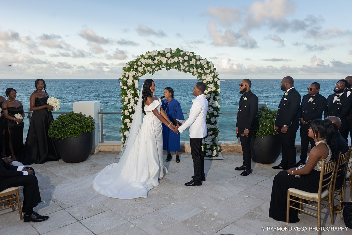 Couple exchanges vows at outdoor oceanfront wedding ceremony with white floral arch, wedding party, and guests on sunny day.