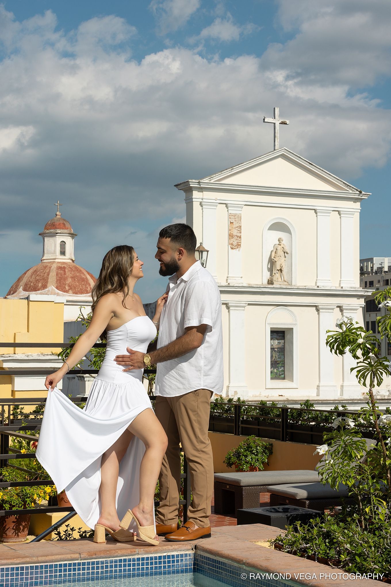Couple in white attire dancing on a rooftop pool terrace with a historic white church and blue sky backdrop.Hotel El convento Old San Juan