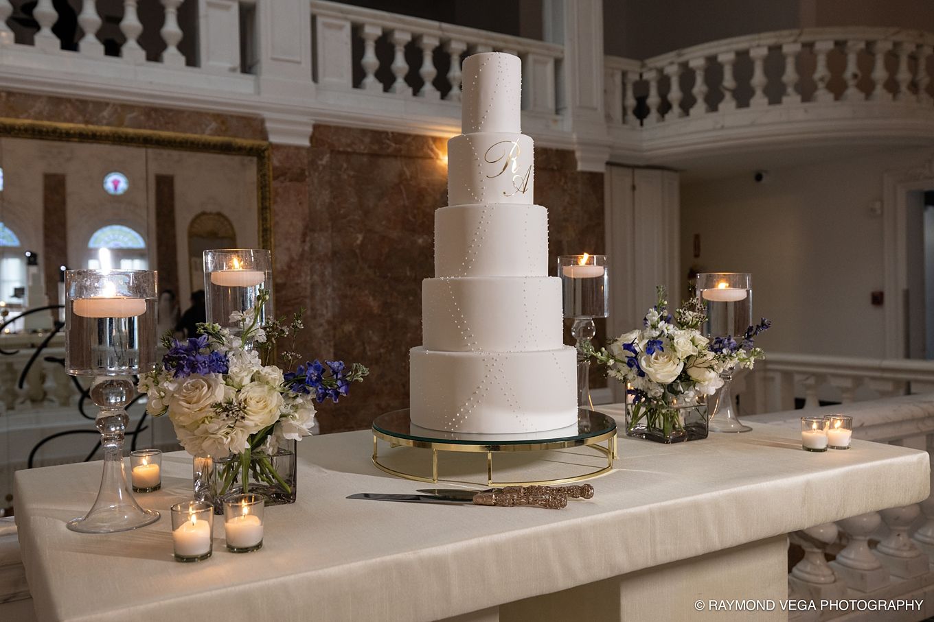 Elegant five-tier white wedding cake on a marble table surrounded by candles and floral arrangements in a grand venue. Antiguo Casino