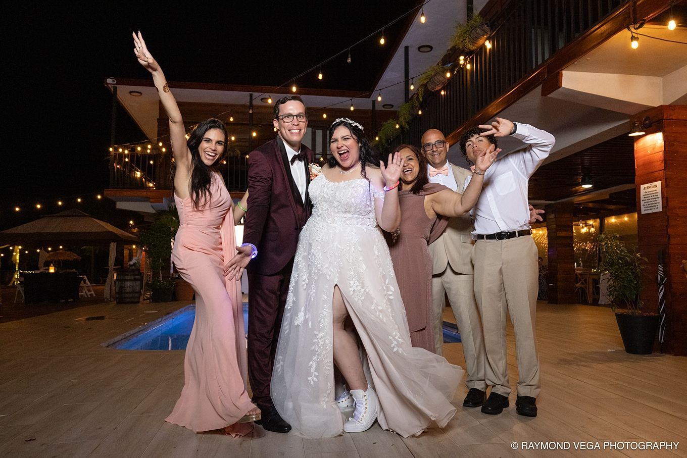 Wedding party celebrates joyfully outdoors at night, bride in white gown and groom in burgundy suit with guests posing.