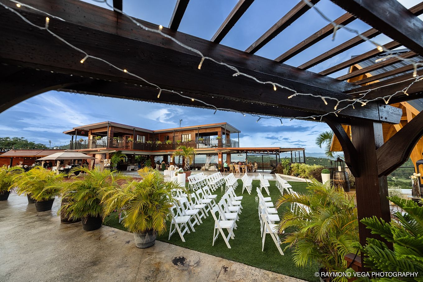 Outdoor wedding venue with white chairs on green lawn, tropical plants, string lights, and a rustic wooden pergola under blue skies.