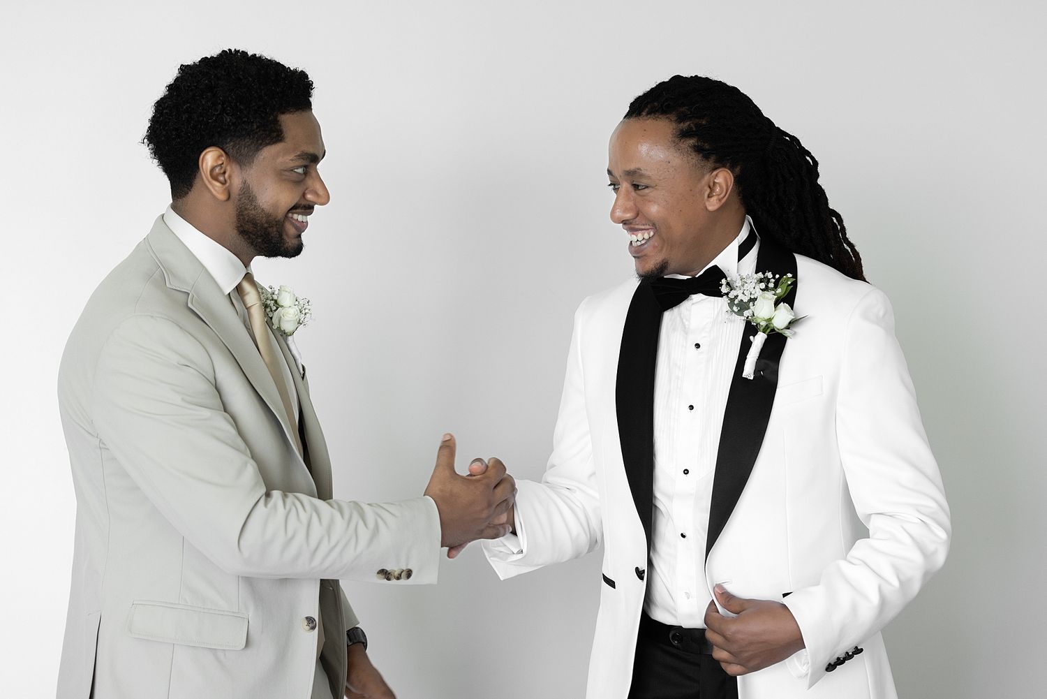 Two well-dressed grooms in formal suits shaking hands and smiling at each other on their wedding day.