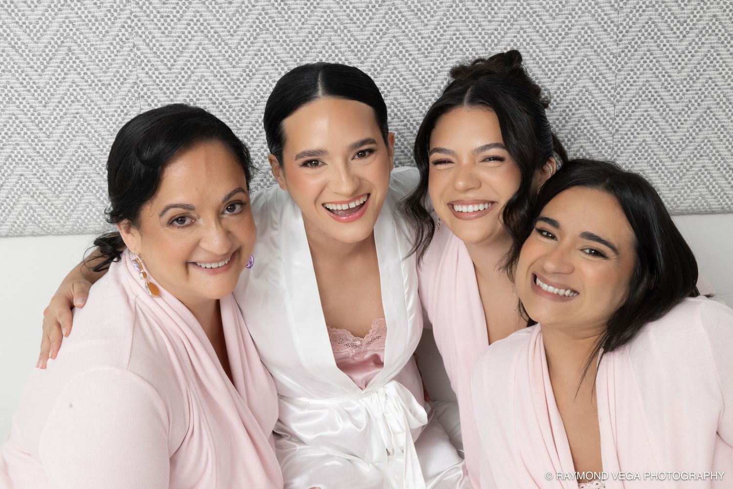 Bride in white satin robe smiling with three bridesmaids in pink robes for a wedding portrait.