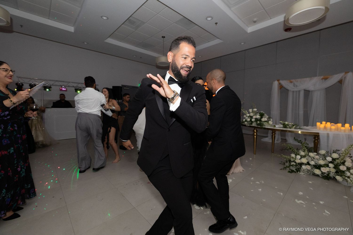 Man in tuxedo dancing joyfully at an elegant wedding reception with guests celebrating in the background.