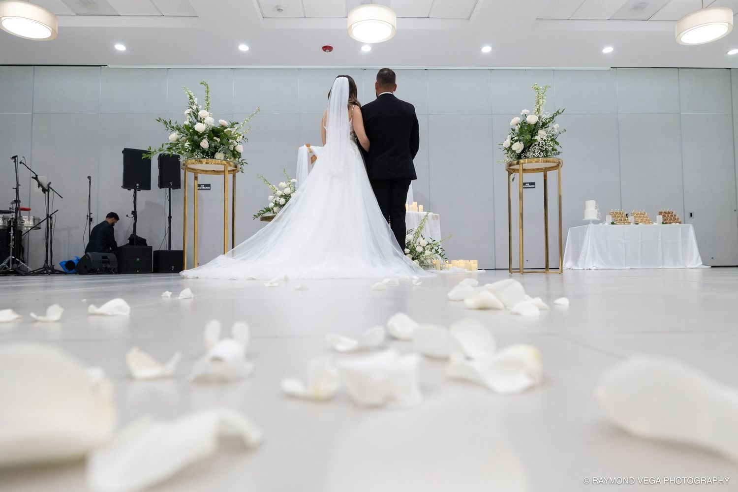 Bride and groom standing at the altar with white rose petals on the floor during an elegant indoor wedding ceremony.