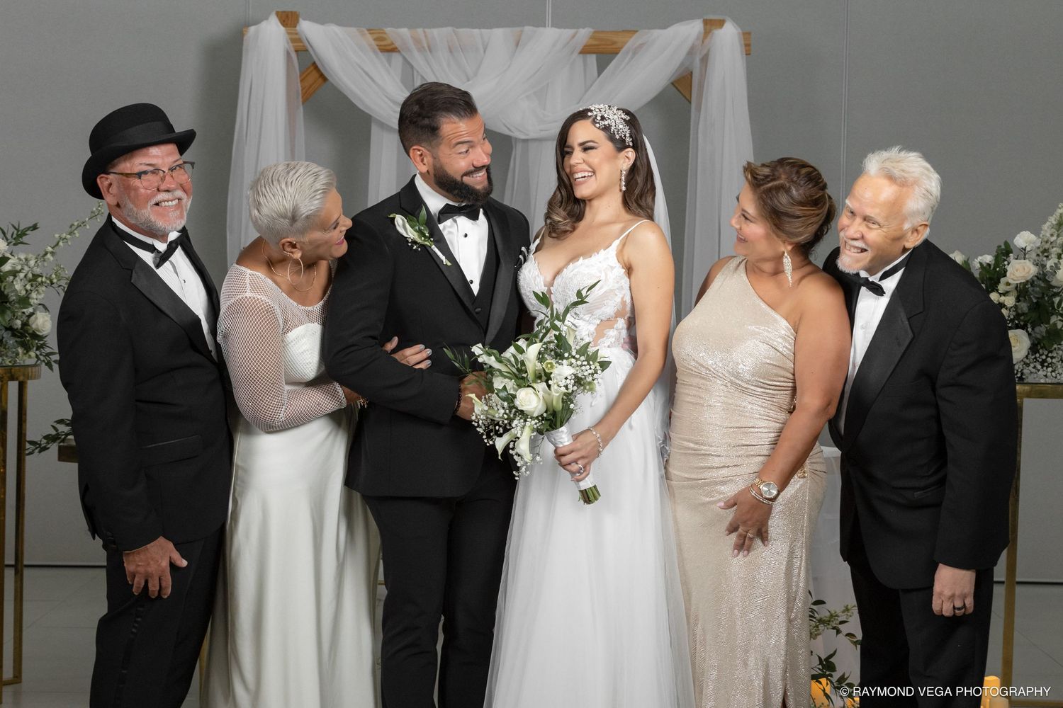 Bride and groom smile together with family members at wedding ceremony under white floral arch.