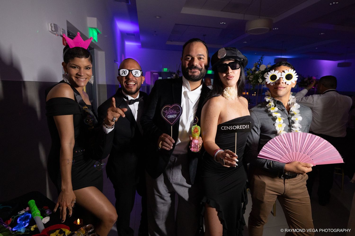 Five friends pose with fun photo booth props at a formal event, wearing black attire and colorful accessories.