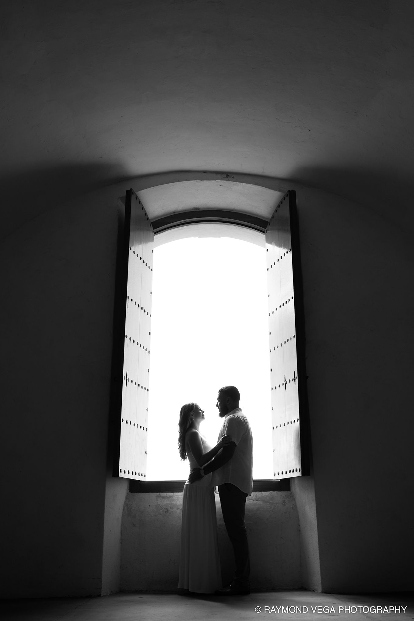 Black and white silhouette of a couple embracing in front of a large arched window, captured by Raymond Vega Photography.