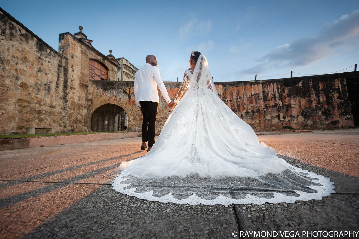 Bride and groom walk hand-in-hand at a historic stone fortress, her lace cathedral veil trailing elegantly on the cobblestone plaza.