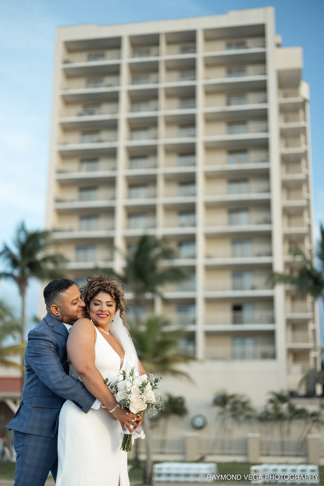 A bride and groom embrace outdoors with a beachfront hotel and palm trees in the background on their wedding day.
