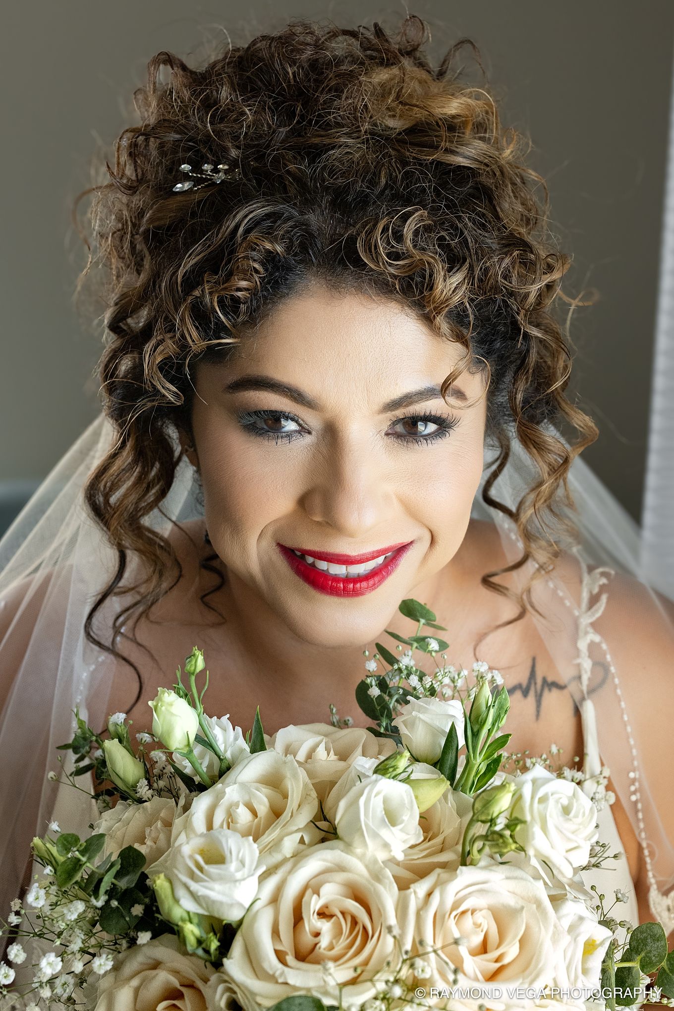 Smiling bride with curly updo, red lips, crystal hair pins, white veil, holding elegant bouquet of cream roses and greenery.