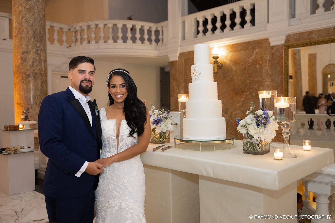 Bride and groom smiling beside a white tiered wedding cake on a marble pedestal in an elegant ballroom venue.Antiguo Casino Puerto Rico