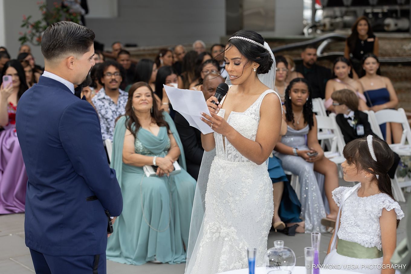 Bride in white gown reads vows at outdoor wedding ceremony while groom and seated guests look on. Antiguo Casino Puerto Rico