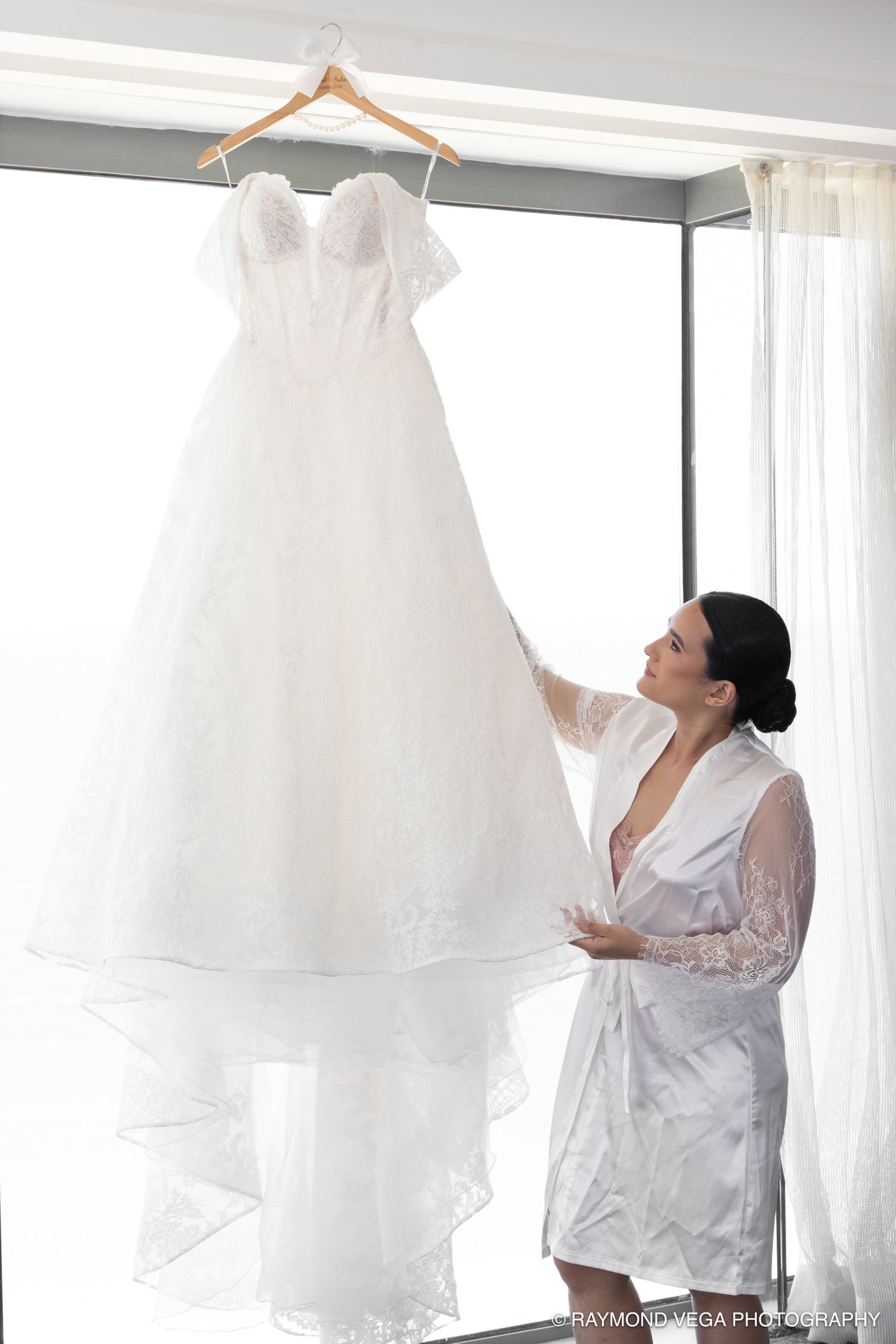 Bride in white robe admiring her elegant lace wedding gown hanging by a bright window before the ceremony.