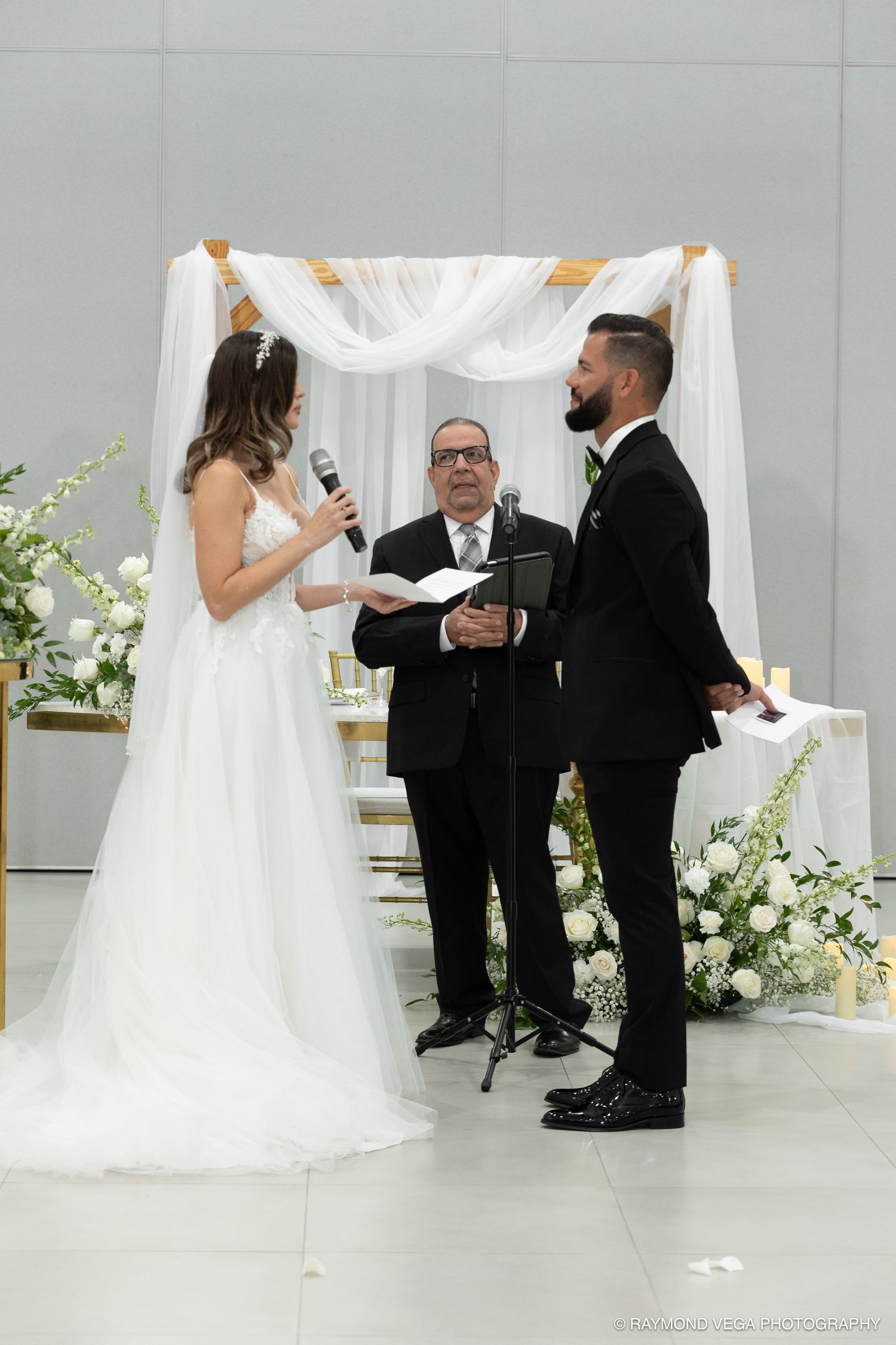 Bride reads vows to groom at elegant indoor wedding ceremony with white floral arch and officiant.
