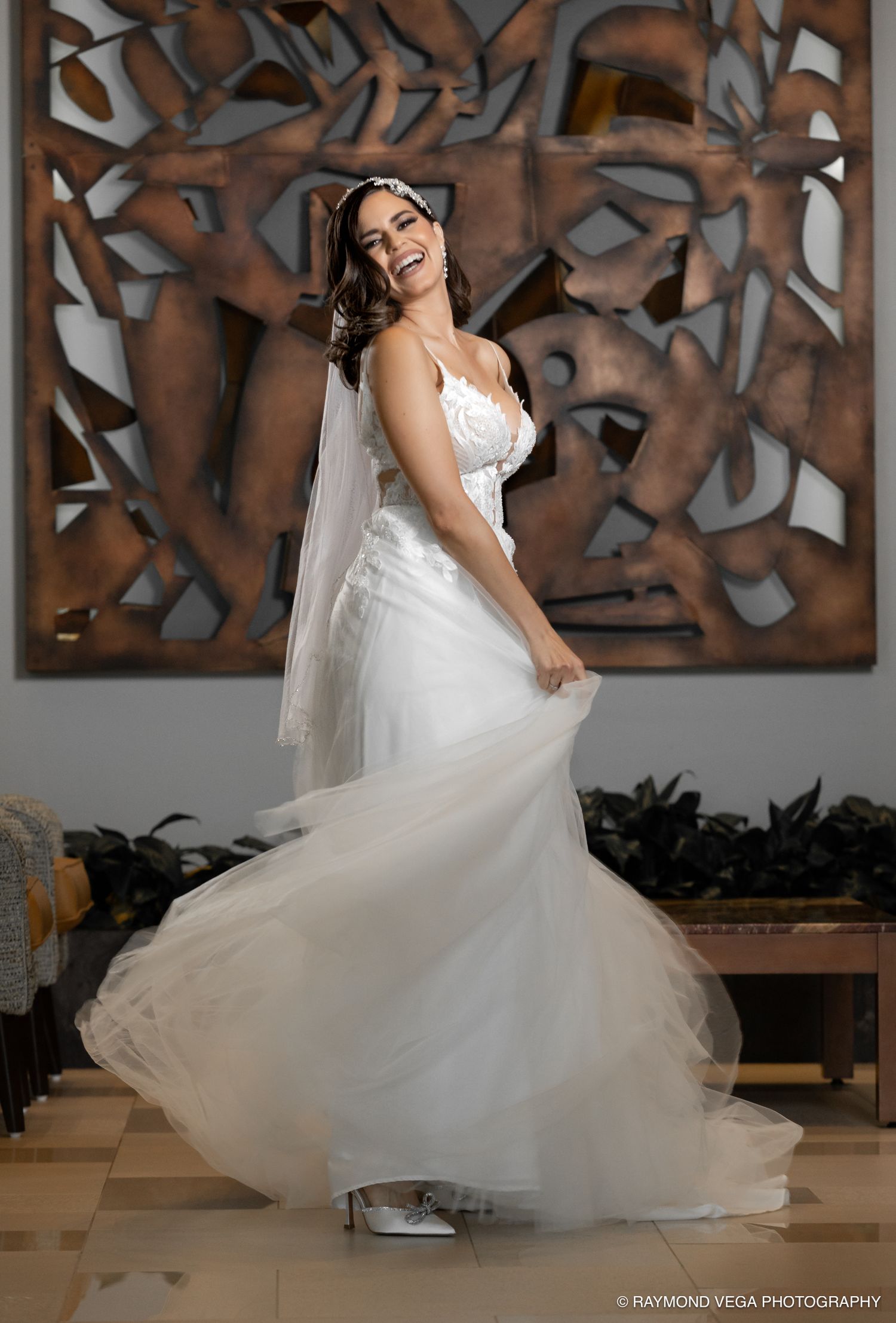 Joyful bride in white gown and veil twirling and laughing in front of decorative wall art.
