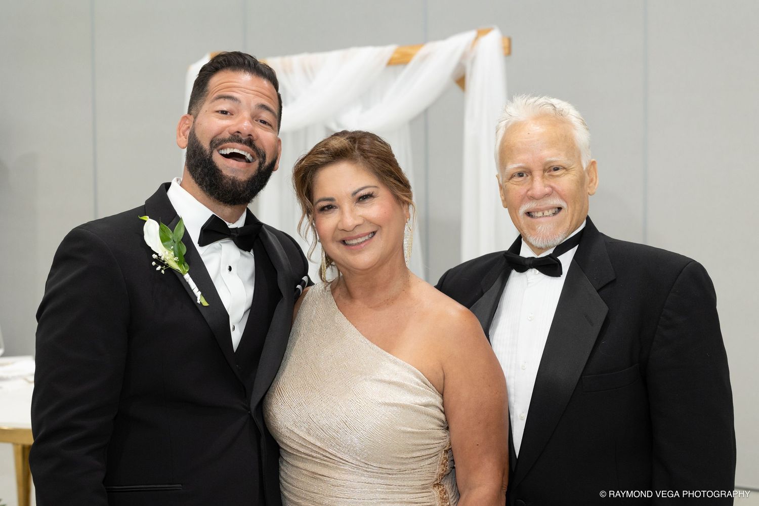 Three people smile at a wedding reception, groom in tuxedo with boutonniere flanked by two guests in formal attire.
