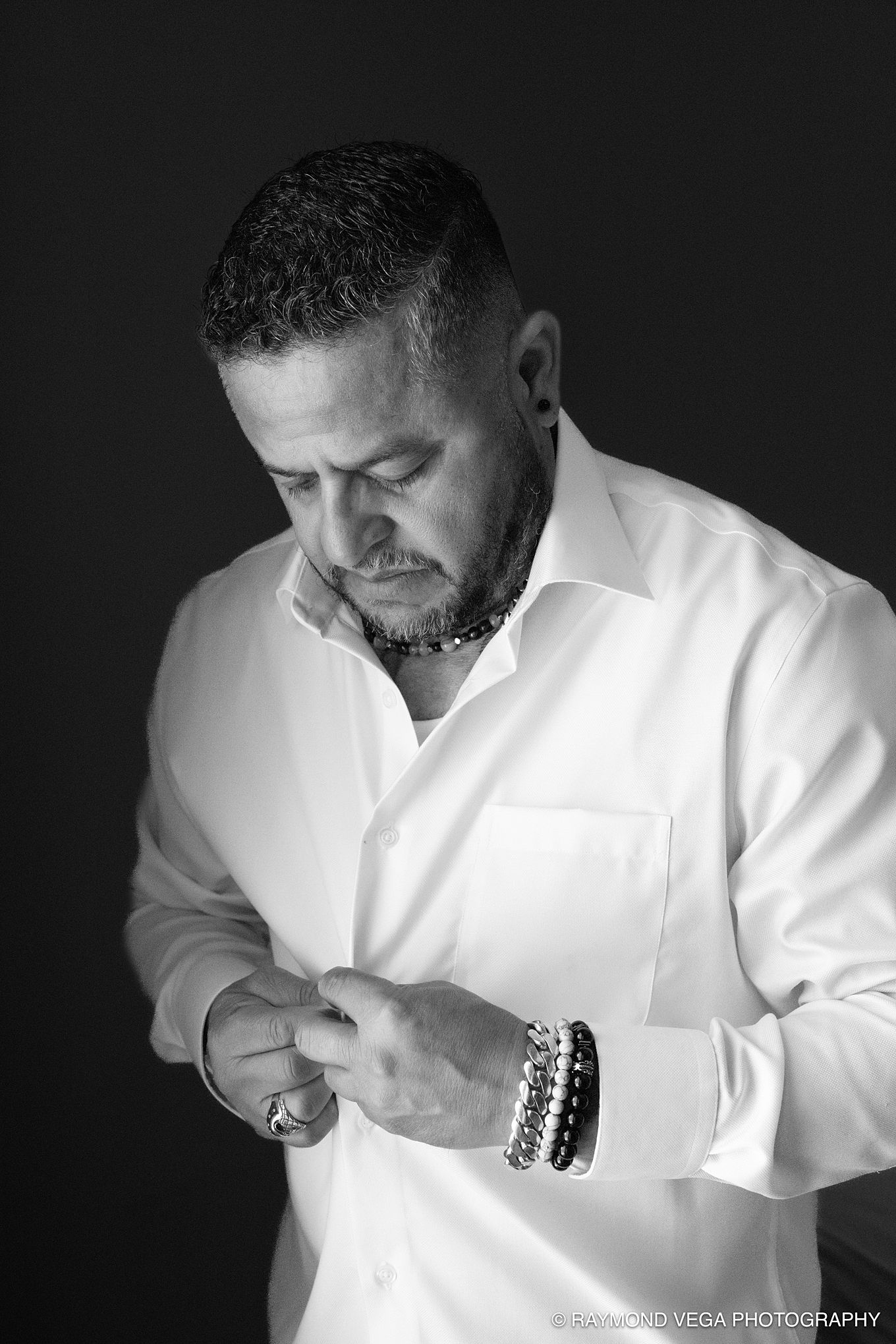 Black and white portrait of a man in a white dress shirt adjusting his cufflinks, wearing bracelets and a ring.