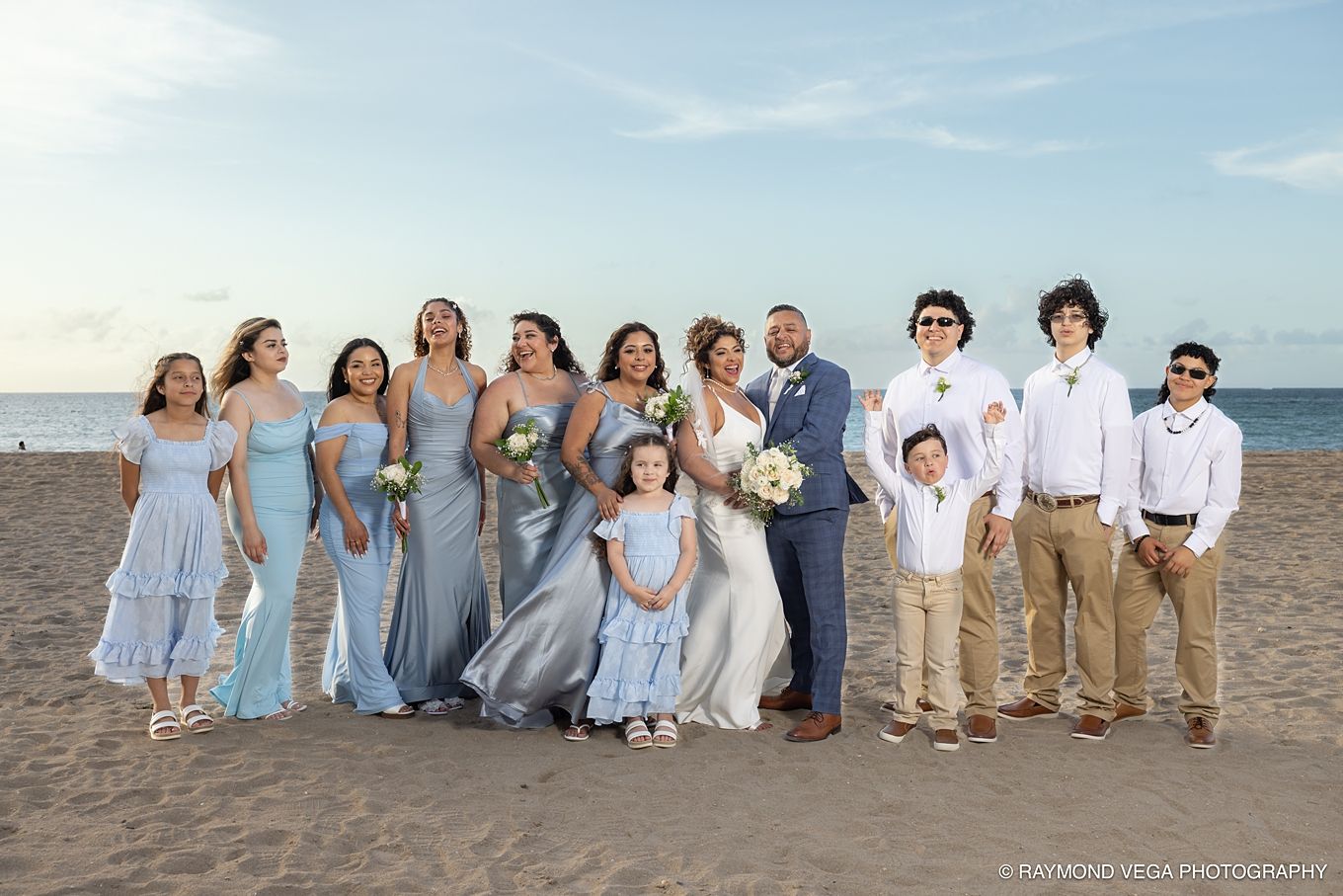 Wedding party poses on beach, bride in silver gown with bridesmaids in blue dresses and groomsmen in khaki pants. at Courtyard by Marriott