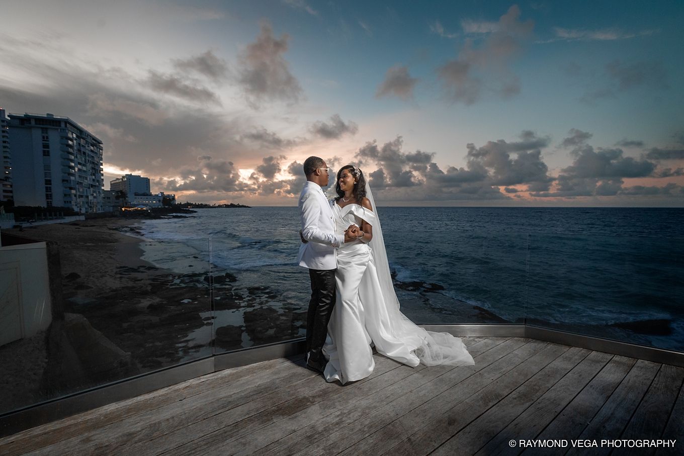 Bride and groom embrace on oceanfront deck at sunset, with dramatic cloudy sky and coastline in background.
