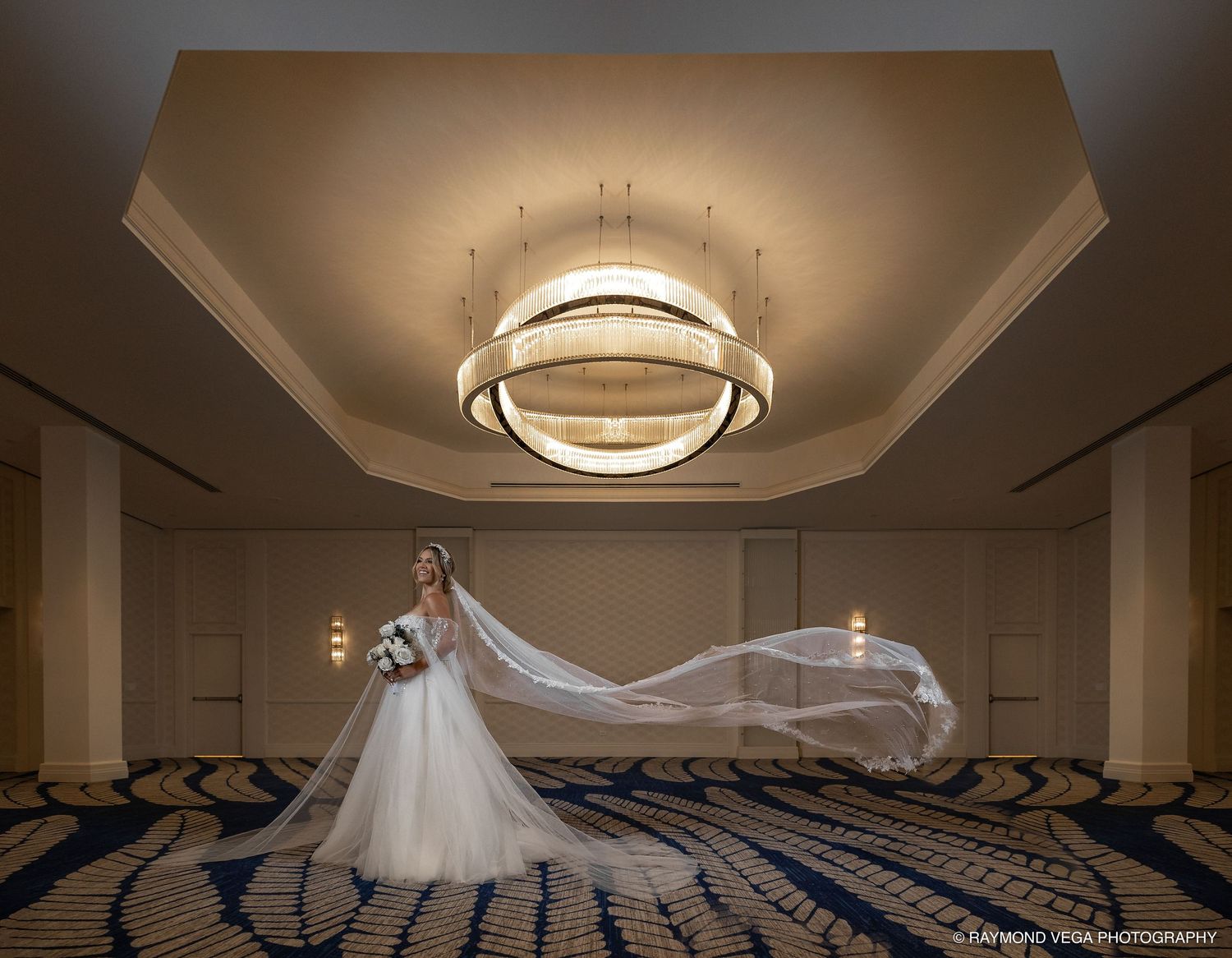 Bride in a flowing wedding gown with long veil billowing under an elegant chandelier in a grand hotel ballroom.