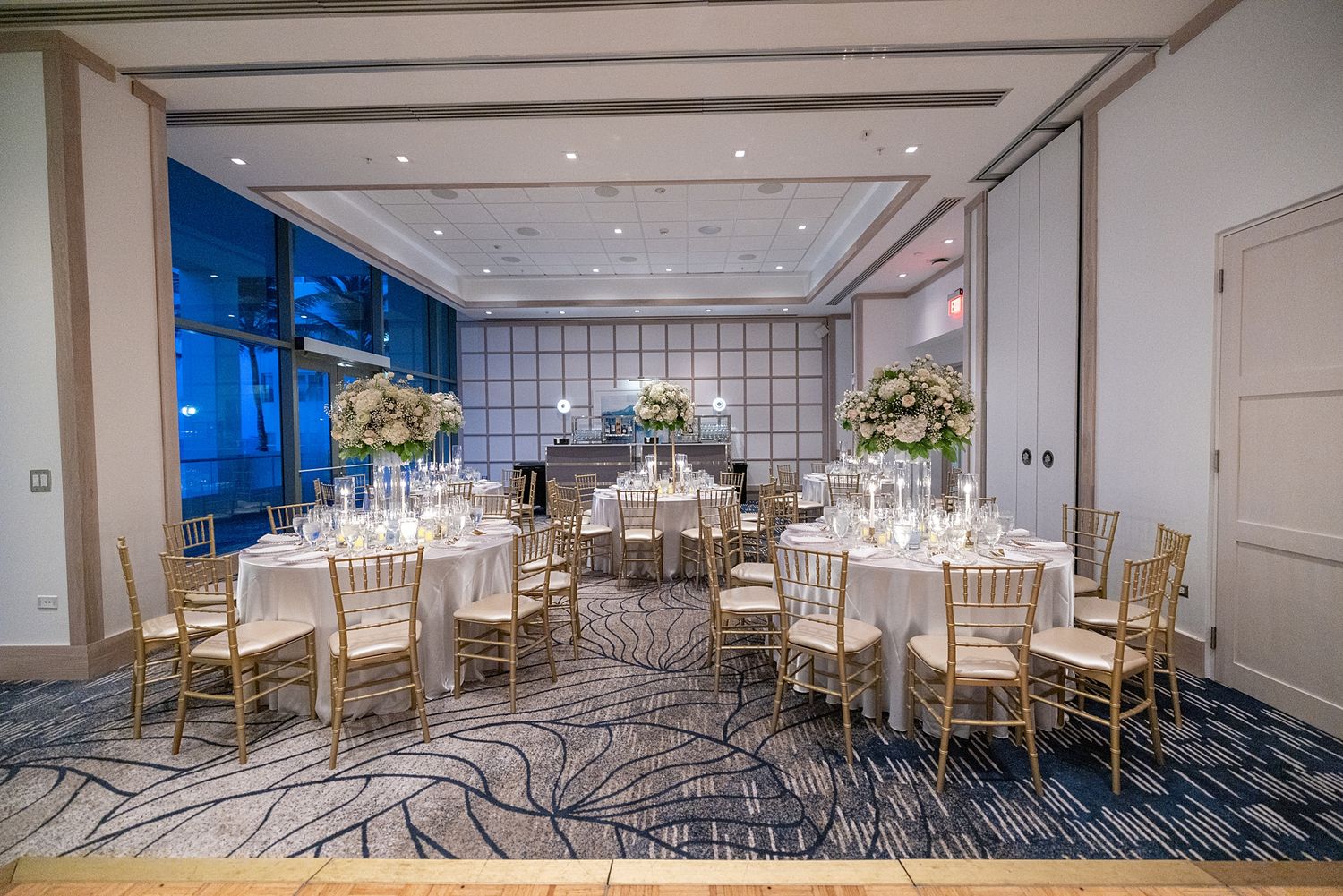 Elegant banquet hall with round tables, gold chiavari chairs, floral centerpieces, and blue patterned carpet for weddings. Reception setup