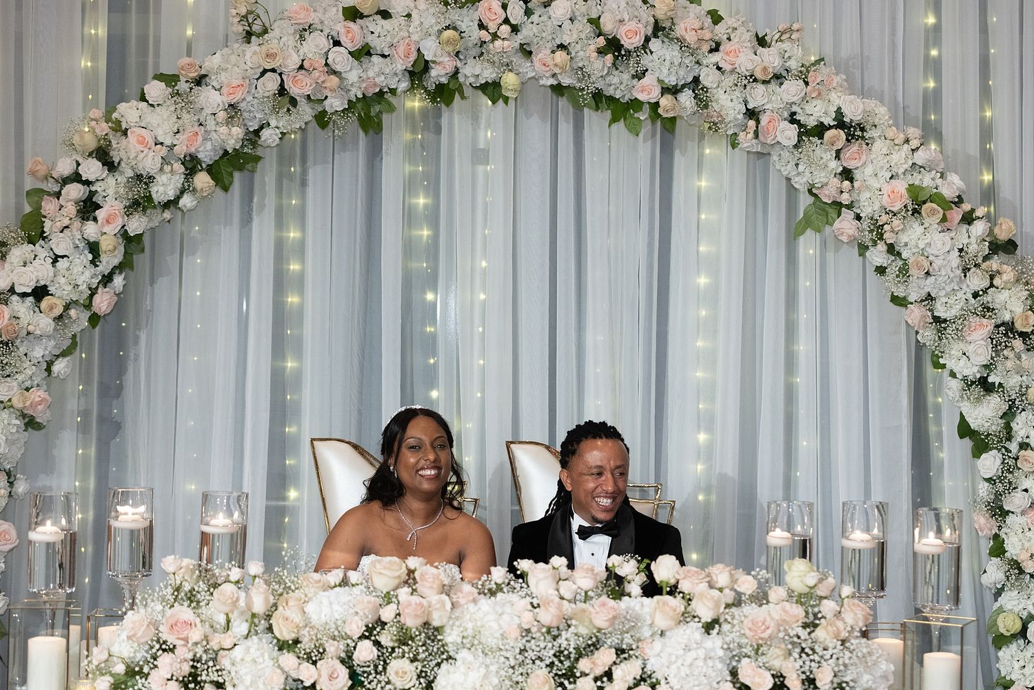 Newlywed couple smiling at sweetheart table under floral arch with pink roses and candles at elegant wedding reception.