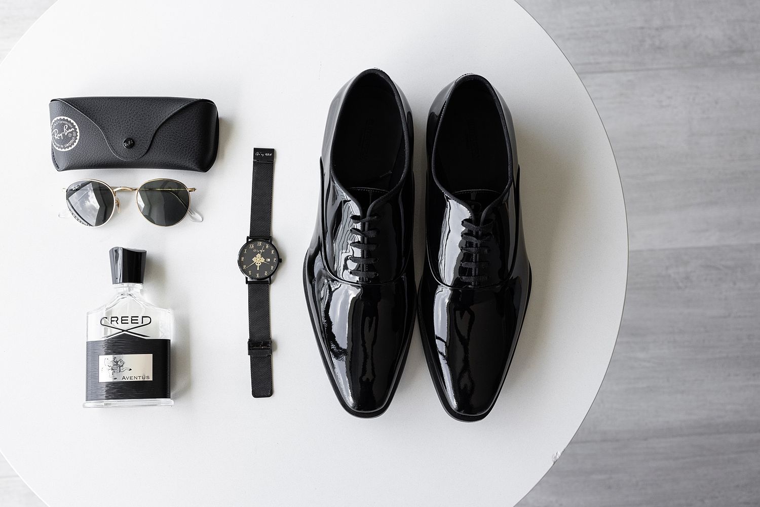 Black patent leather Oxford shoes displayed with Ray-Ban sunglasses, Creed cologne, and a black watch on a white table.