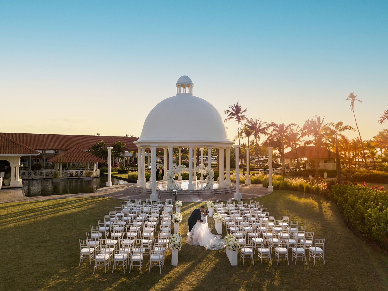 Aerial view of an elegant outdoor wedding ceremony at a tropical resort with a white gazebo and palm trees at sunset. Hyatt