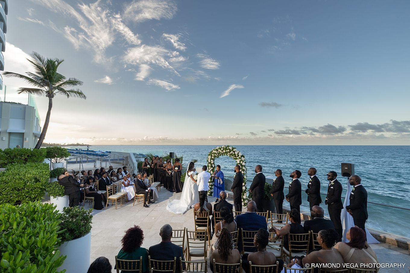 Outdoor beachside wedding ceremony at sunset with ocean views, floral arch, and guests seated on a luxury resort terrace.
