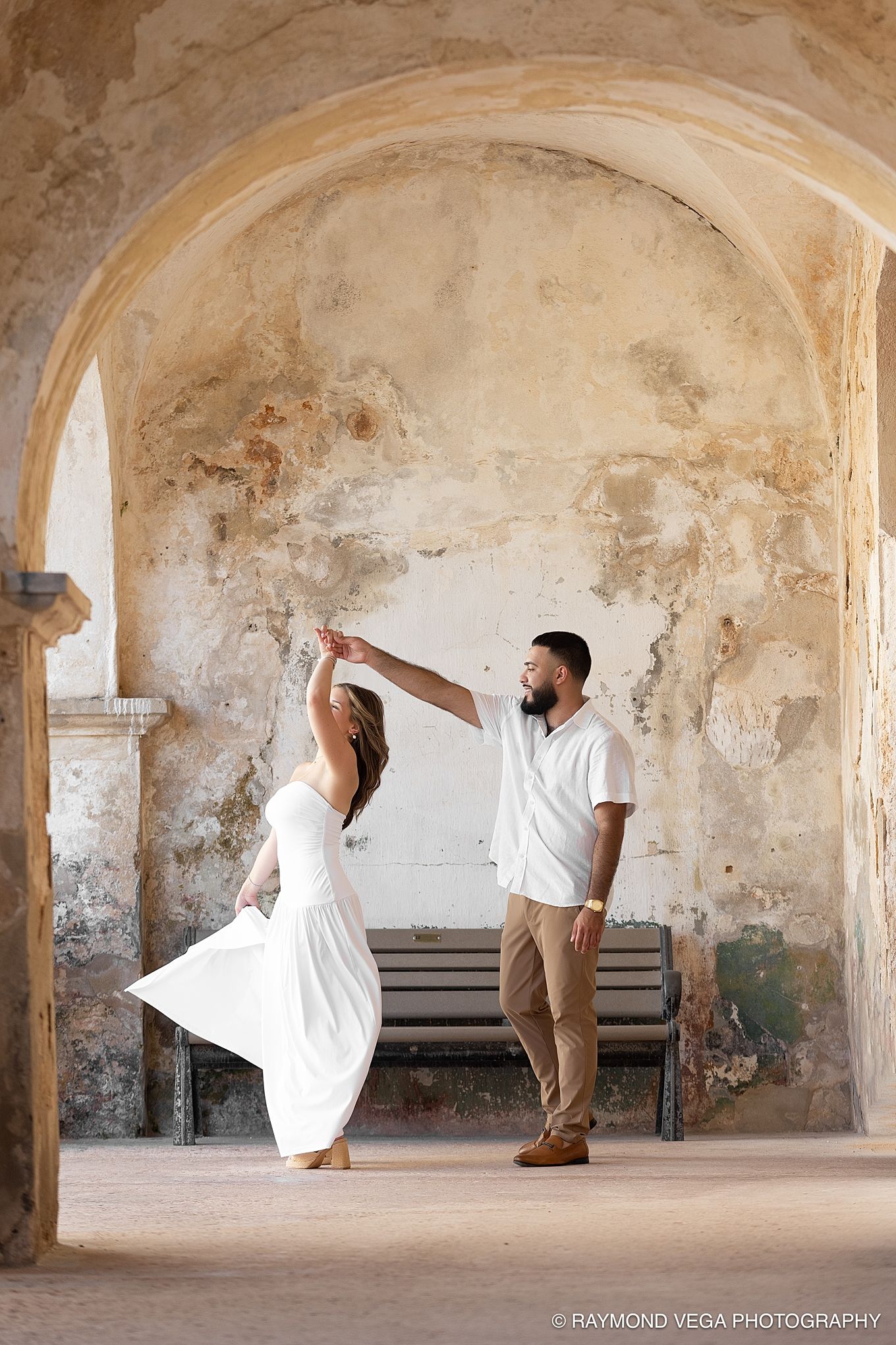 Couple dancing in white outfits under a rustic stone arch at a historic venue, captured by Raymond Vega Photography.