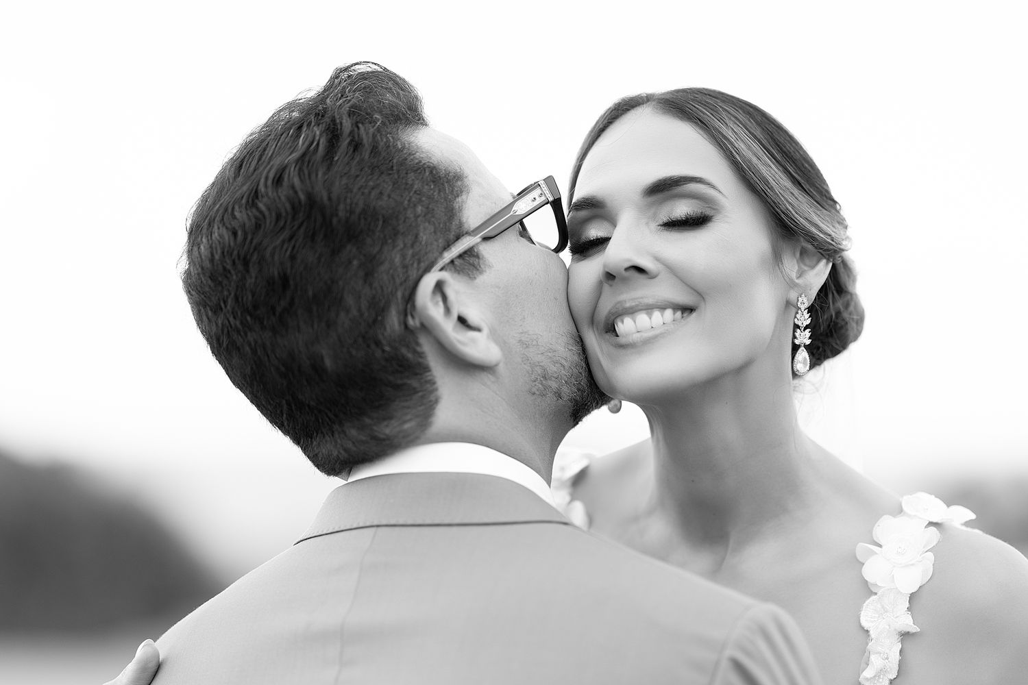 Black and white photo of a groom kissing his bride on the cheek as she smiles joyfully on their wedding day. Raymond Vega Photography