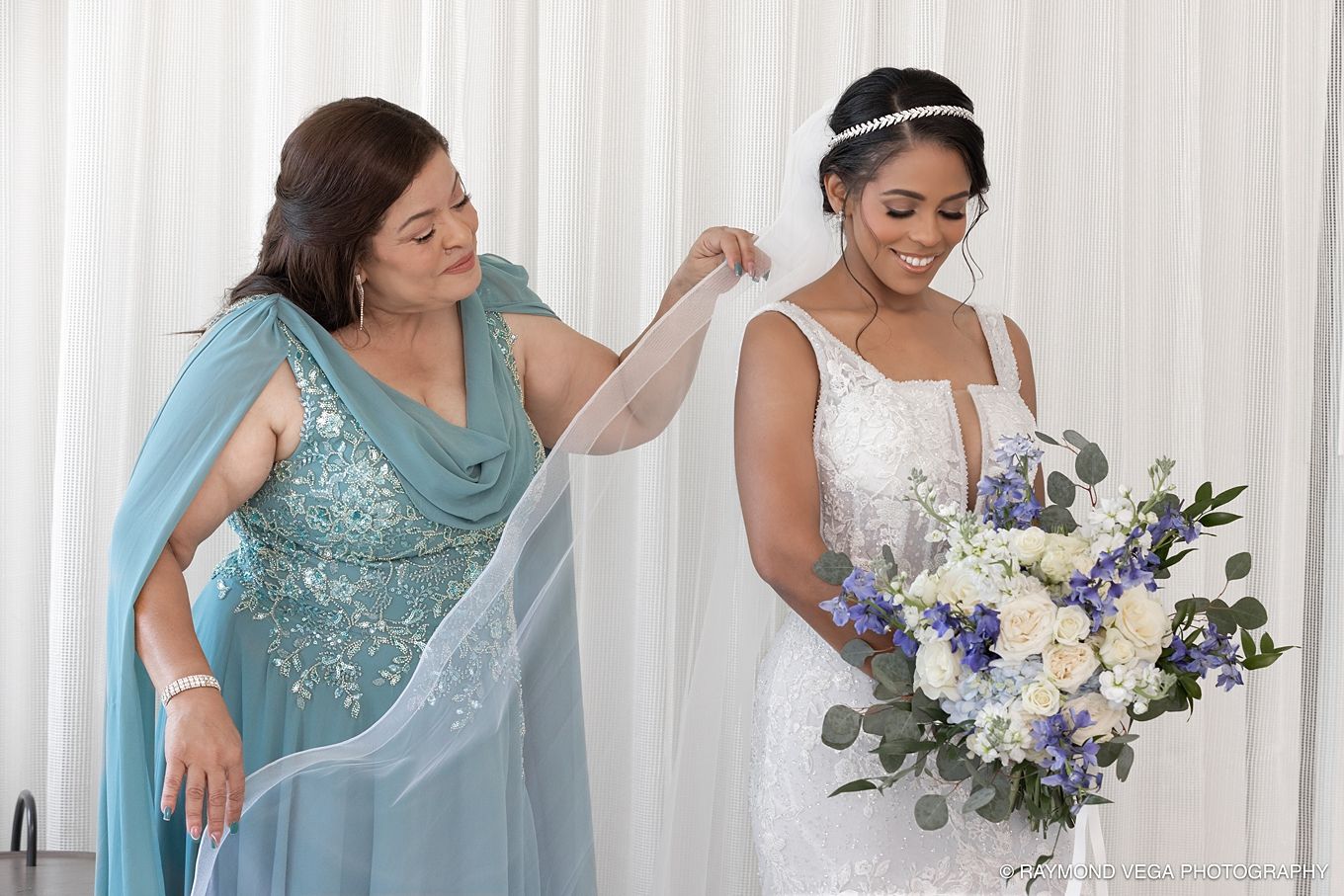 Bride in white lace gown holding blue floral bouquet while woman in teal gown adjusts her veil indoors. Hotel la Concha Wedding